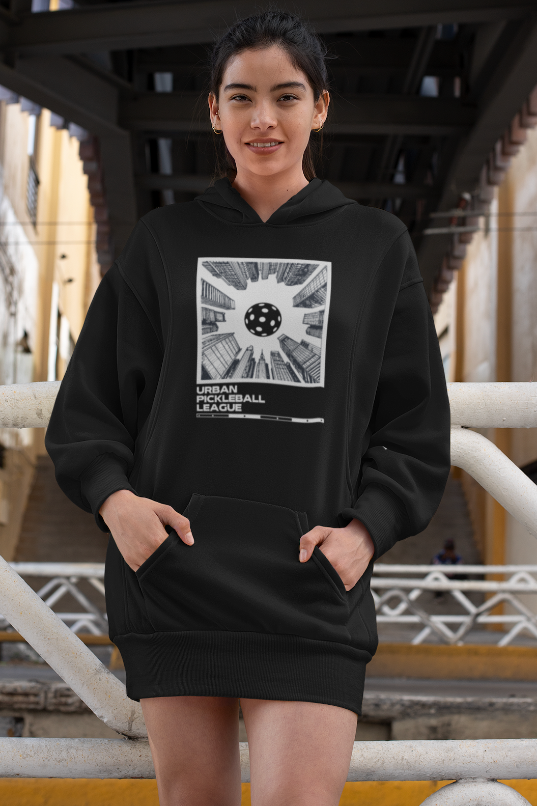Person wearing a black hoodie with a graphic design under a bridge.