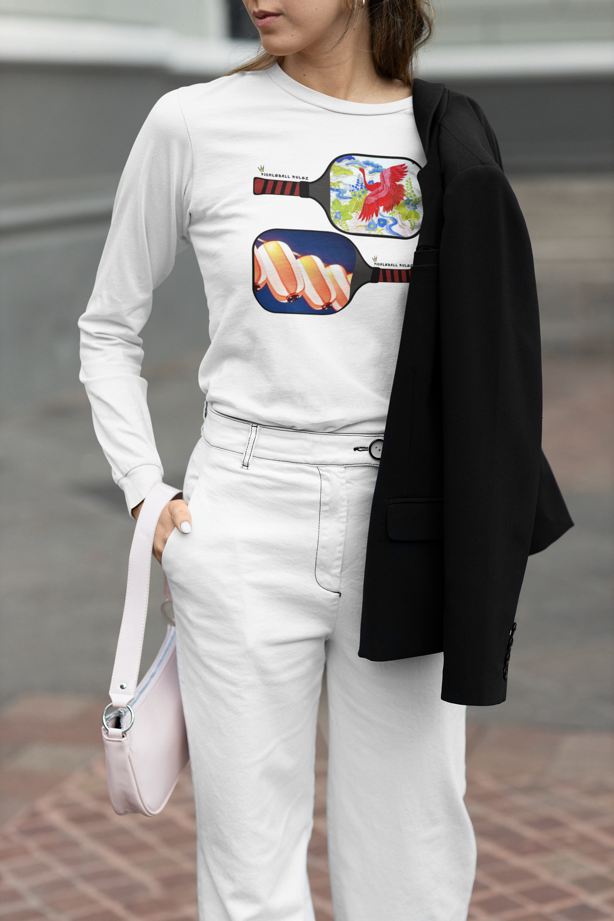 Person wearing a white long-sleeve shirt with colorful pickleball paddle designs and white pants, holding a pink handbag.