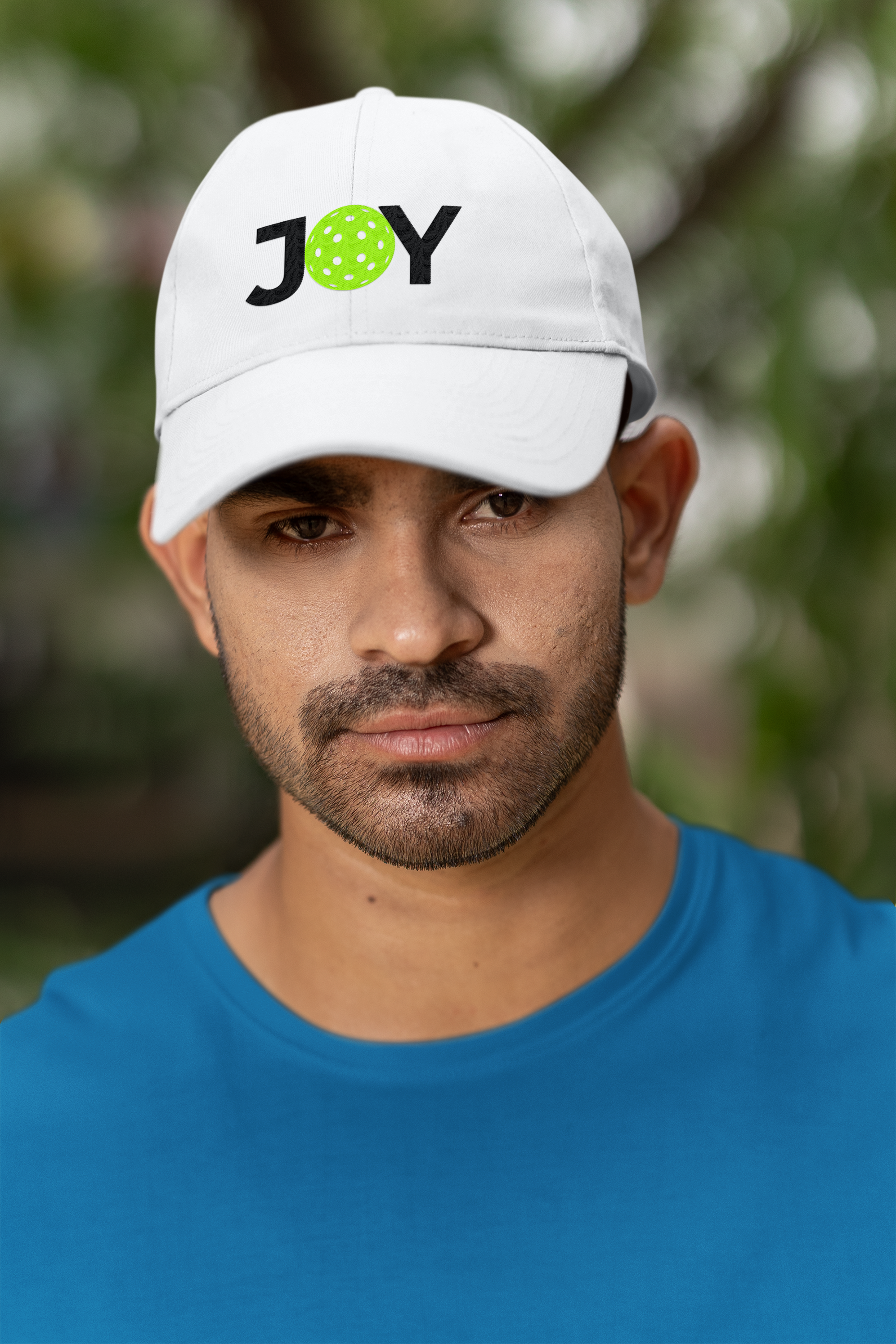 Man wearing a white cap with 'JOY' and a pickleball ball design, outdoors.