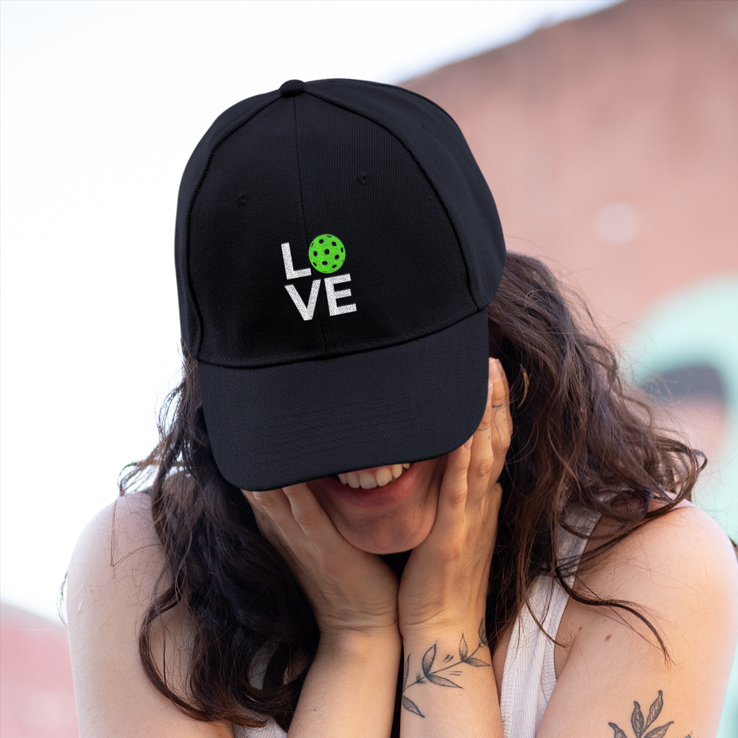 Person wearing a black cap with a logo, white shirt, and dark jeans, standing in front of a blurred background.