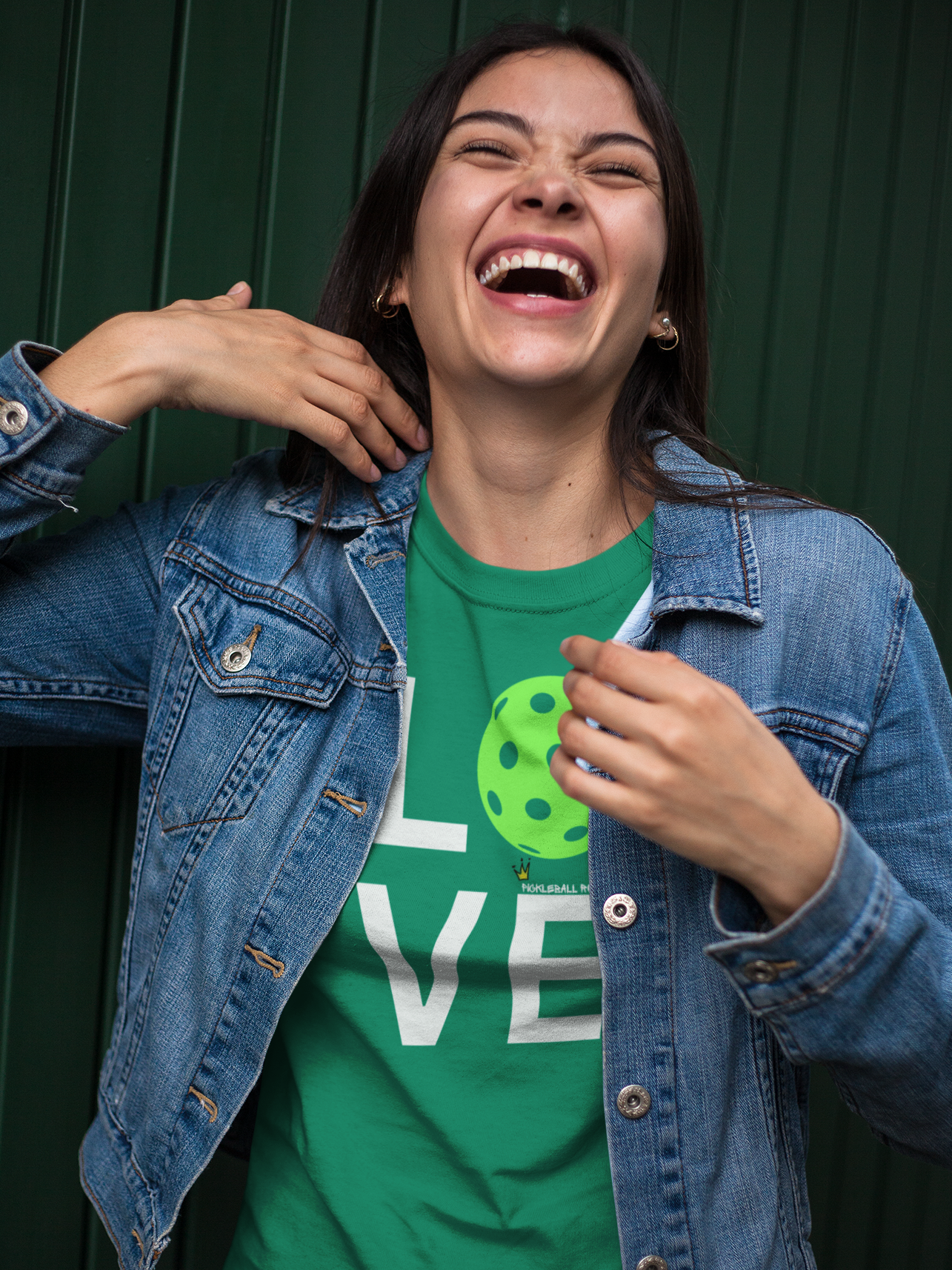 Person wearing a green t-shirt with a logo and a denim jacket, standing against a dark background
