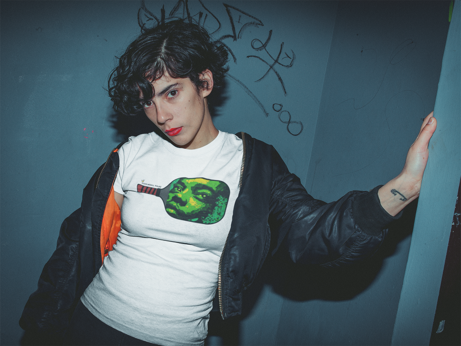 Person wearing a white t-shirt with a graphic design, standing against a blue wall with graffiti.