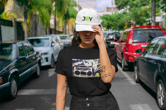 Person wearing a black t-shirt with a graphic design and a white cap with 'JOY' text on a street.