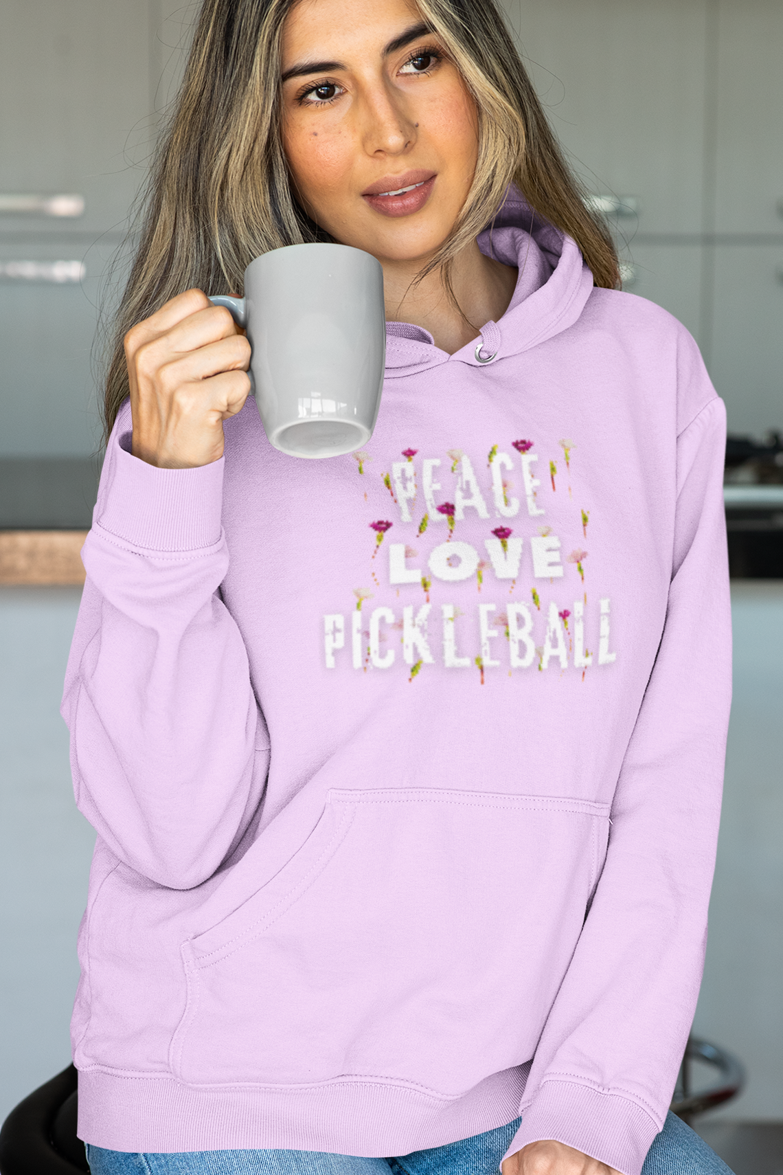 Woman wearing a purple 'Peace Love Pickleball' hoodie holding a mug in a kitchen.