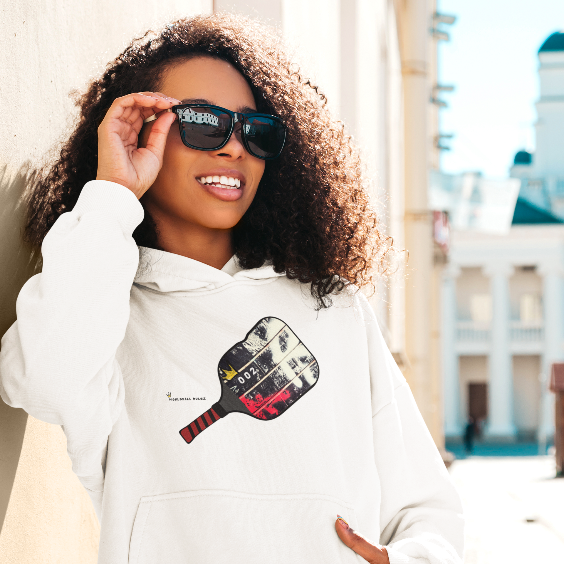 Woman wearing a white hoodie with a graphic design, holding sunglasses, outdoors.