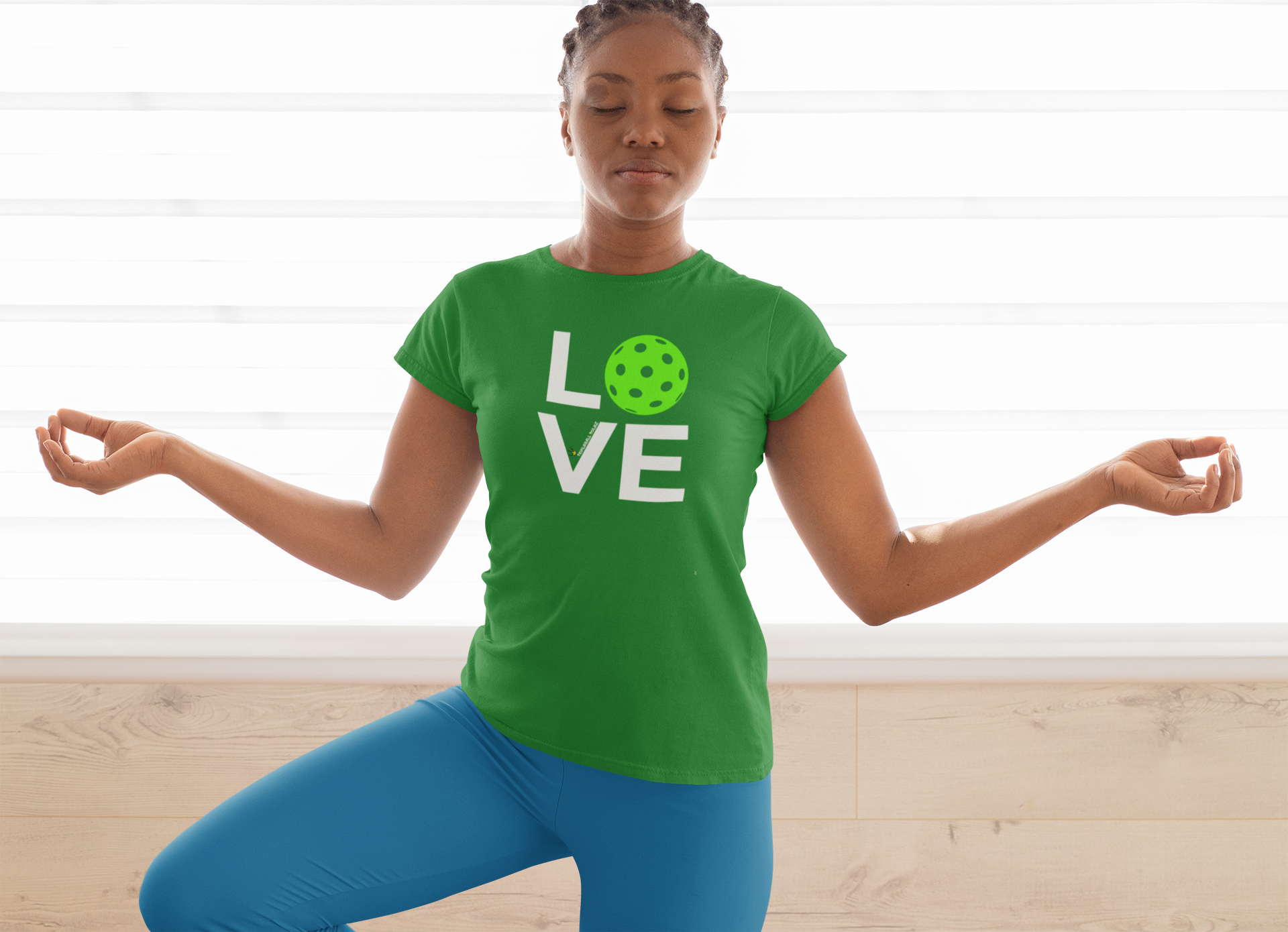 Person wearing a green 'LOVE' t-shirt with a pickleball graphic, meditating indoors.