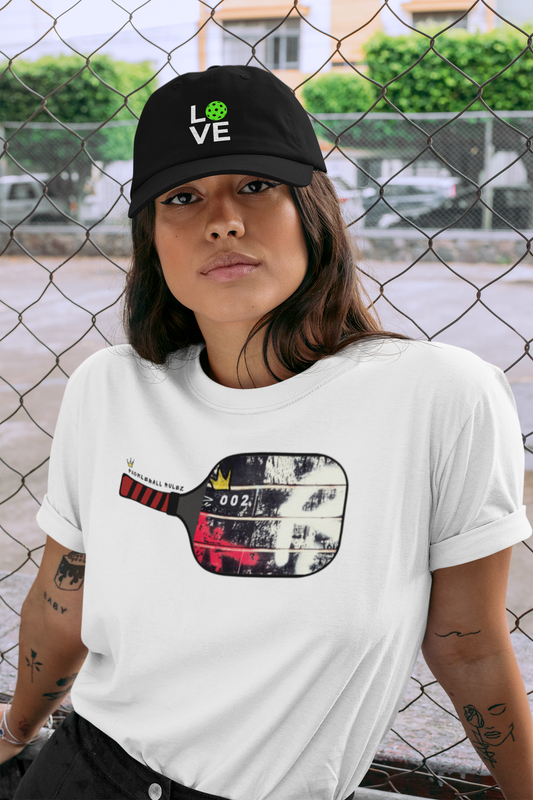 Person wearing a white t-shirt with a graphic design and a black cap with a logo, standing in front of a chain-link fence.