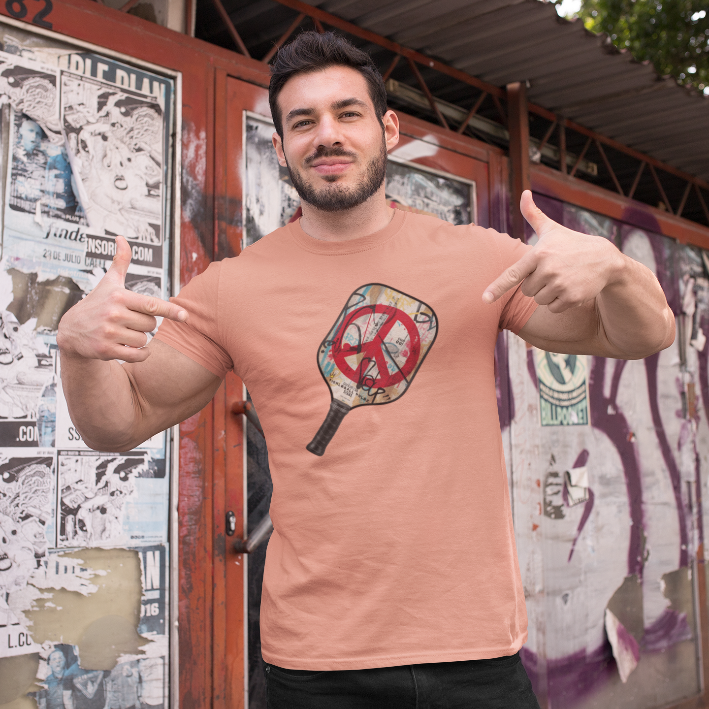 Man wearing a peach t-shirt with a graphic design, giving a thumbs-up gesture in front of a wall with posters.