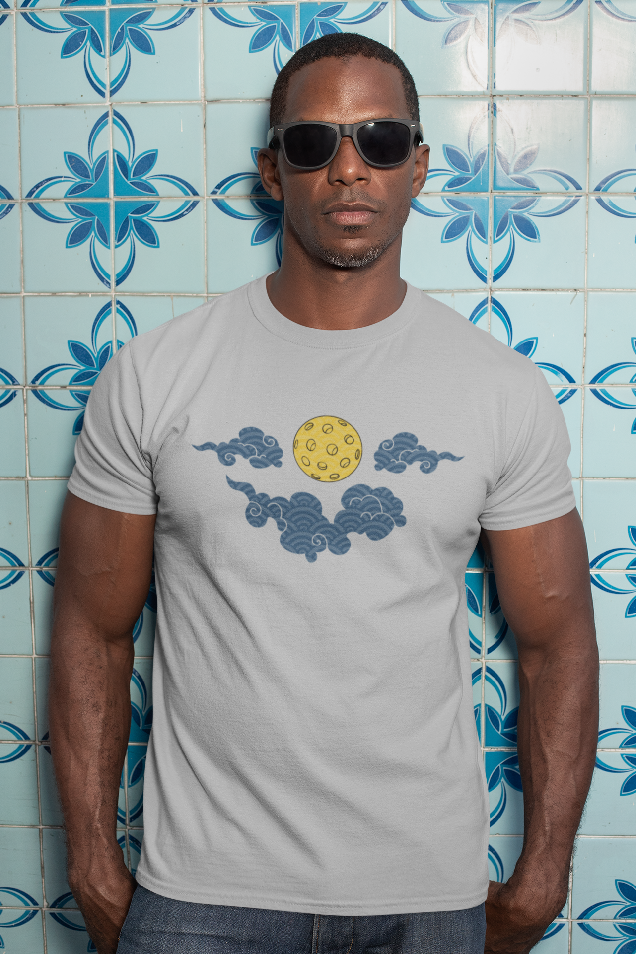 Man wearing a gray t-shirt with a yellow circle and blue cloud design against a blue tiled wall.