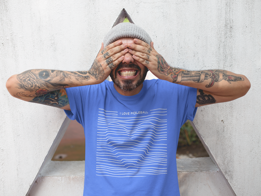 Man with tattoos wearing a blue t-shirt with text, covering his face against a light-colored wall.