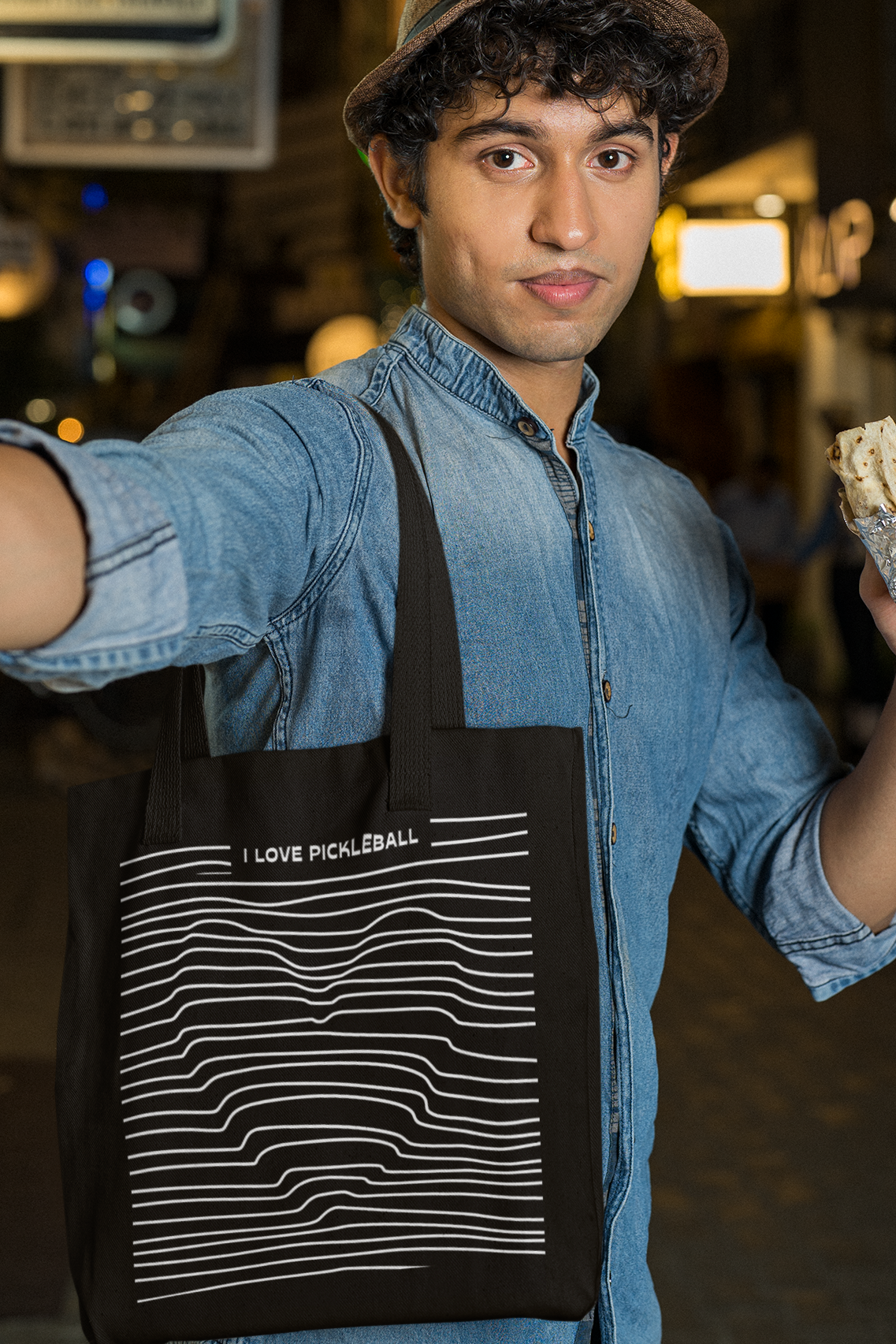 Person wearing a black apron with white text, holding a bagel and taking a selfie in an urban setting.