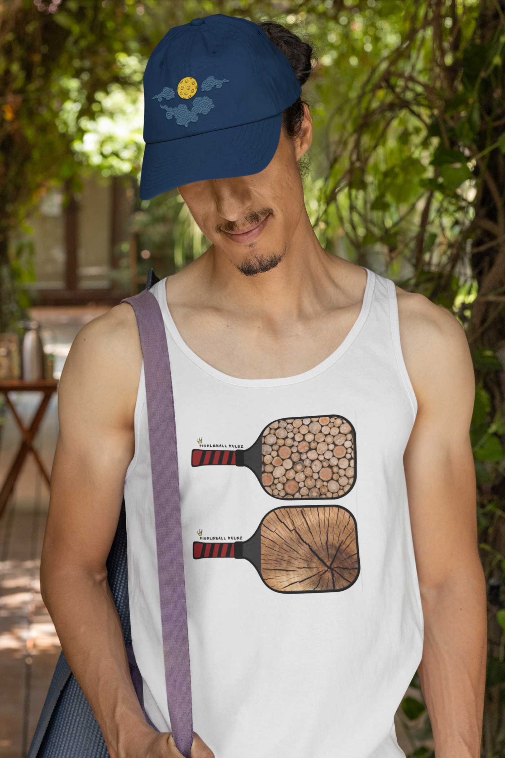 Man wearing a white tank top with paddle ball designs and a blue cap, standing outdoors.